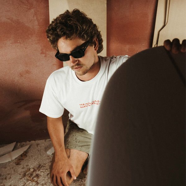 Man bending down and admiring a surfboard in a shaping bay wearing black sunglasses